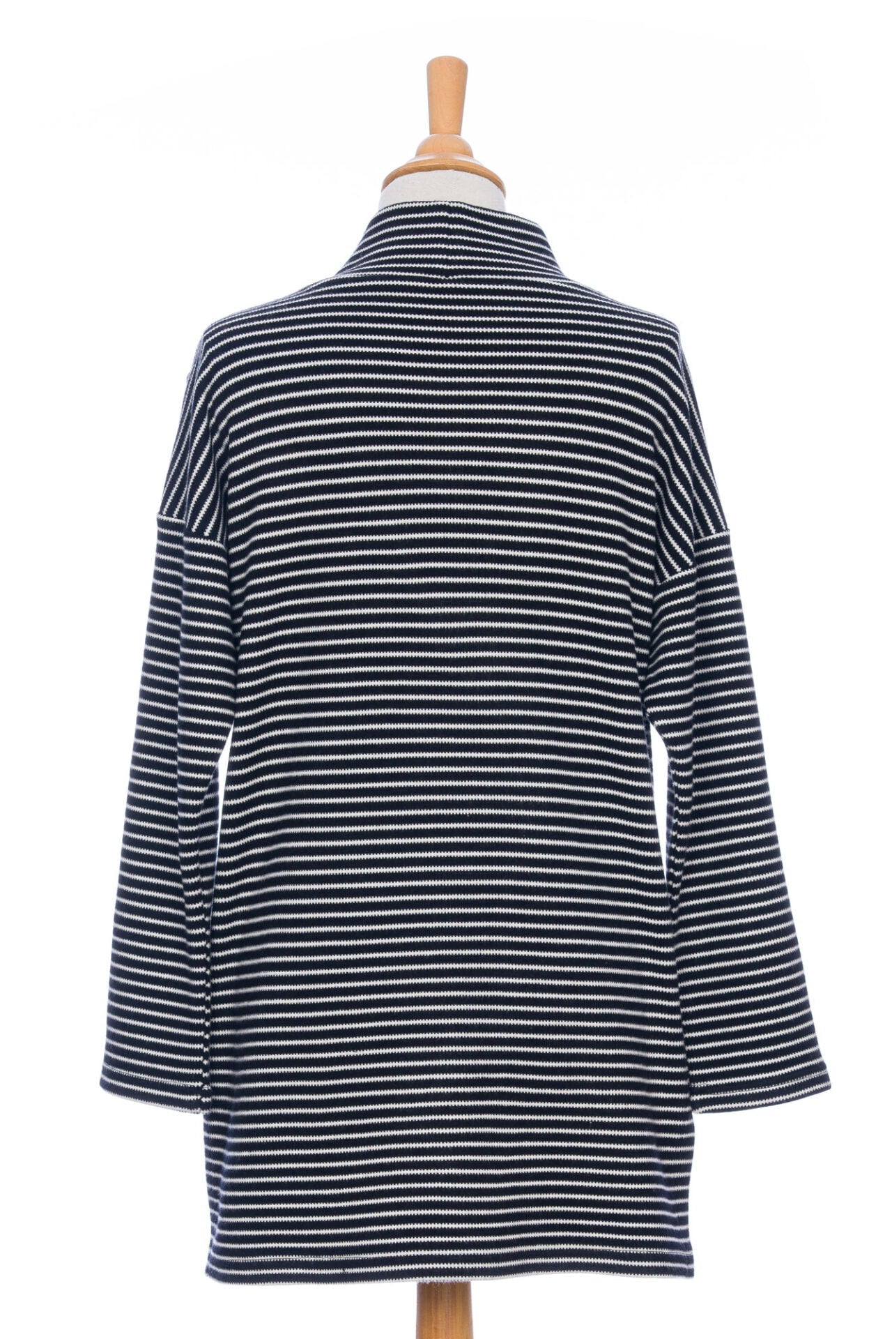 A back view of the Aralie Tunic by Rien ne se Perd in Black and White Stripe, with a high neckline, long sleeves, side slits for ease, drop sleeves and a coconut button placket on the left shoulder, is shown on a mannequin against a white background. 
