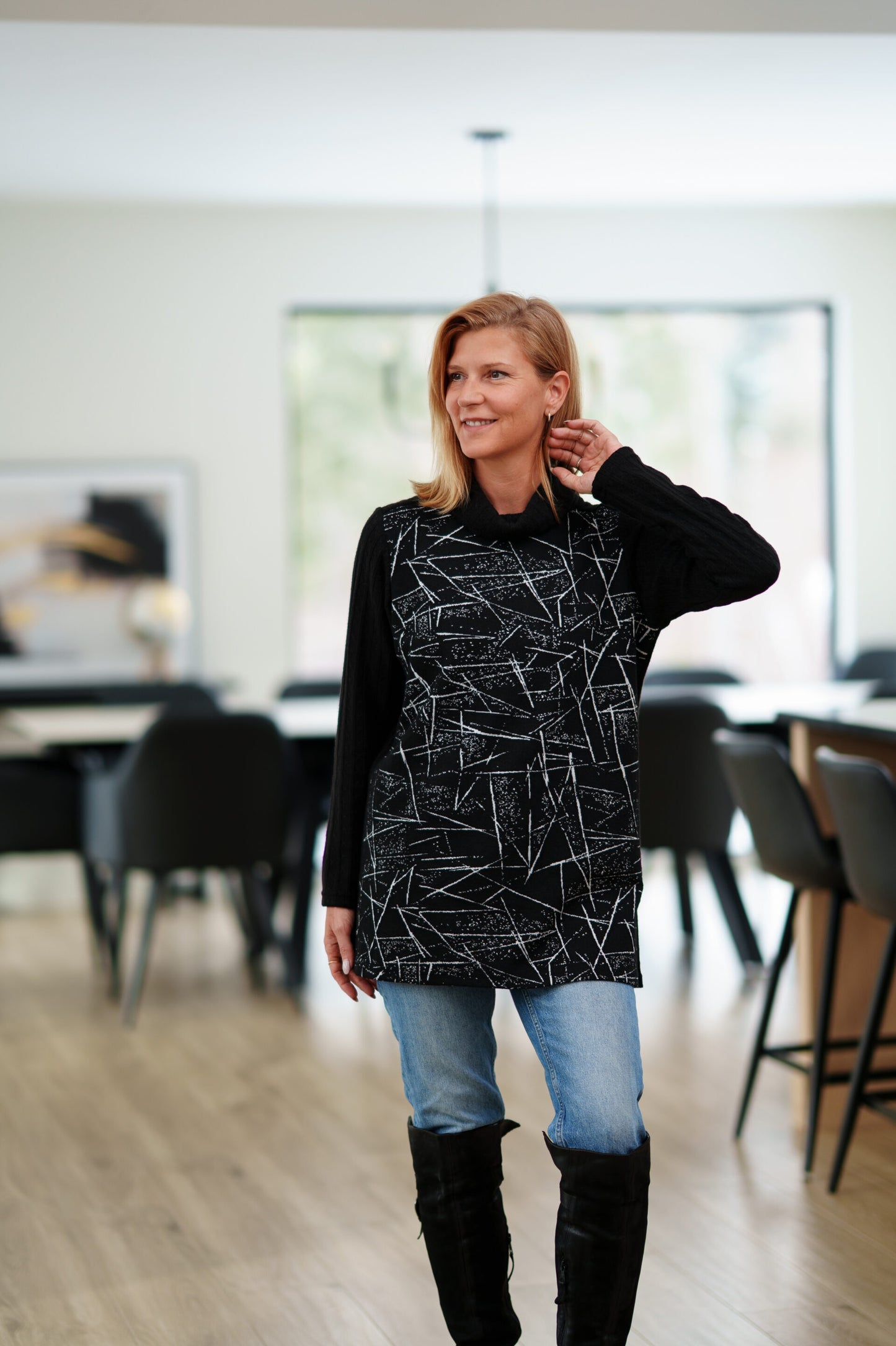 A woman wearing the Auclair Tunic by Rien ne se Perd in a black and white geometric print, with a solid black turtleneck, back, and sleeves, and side slits. She is wearing it with jeans and standing in a room with indoors with a dining room table behind her. 