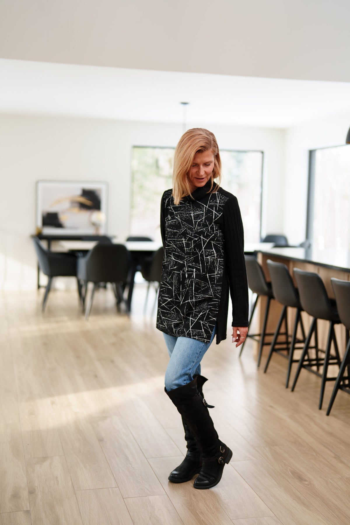 A woman wearing the Auclair Tunic by Rien ne se Perd in a black and white geometric print, with a solid black turtleneck, back, and sleeves, and side slits. She is wearing it with jeans and standing in a room with indoors with a dining room table behind her. 