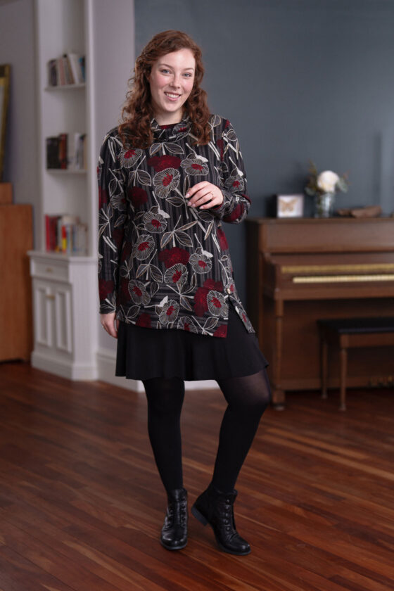 A woman wearing the Corno Tunic by Rien ne se Perd in Black and Red with a black skirt stands in front of a piano. 