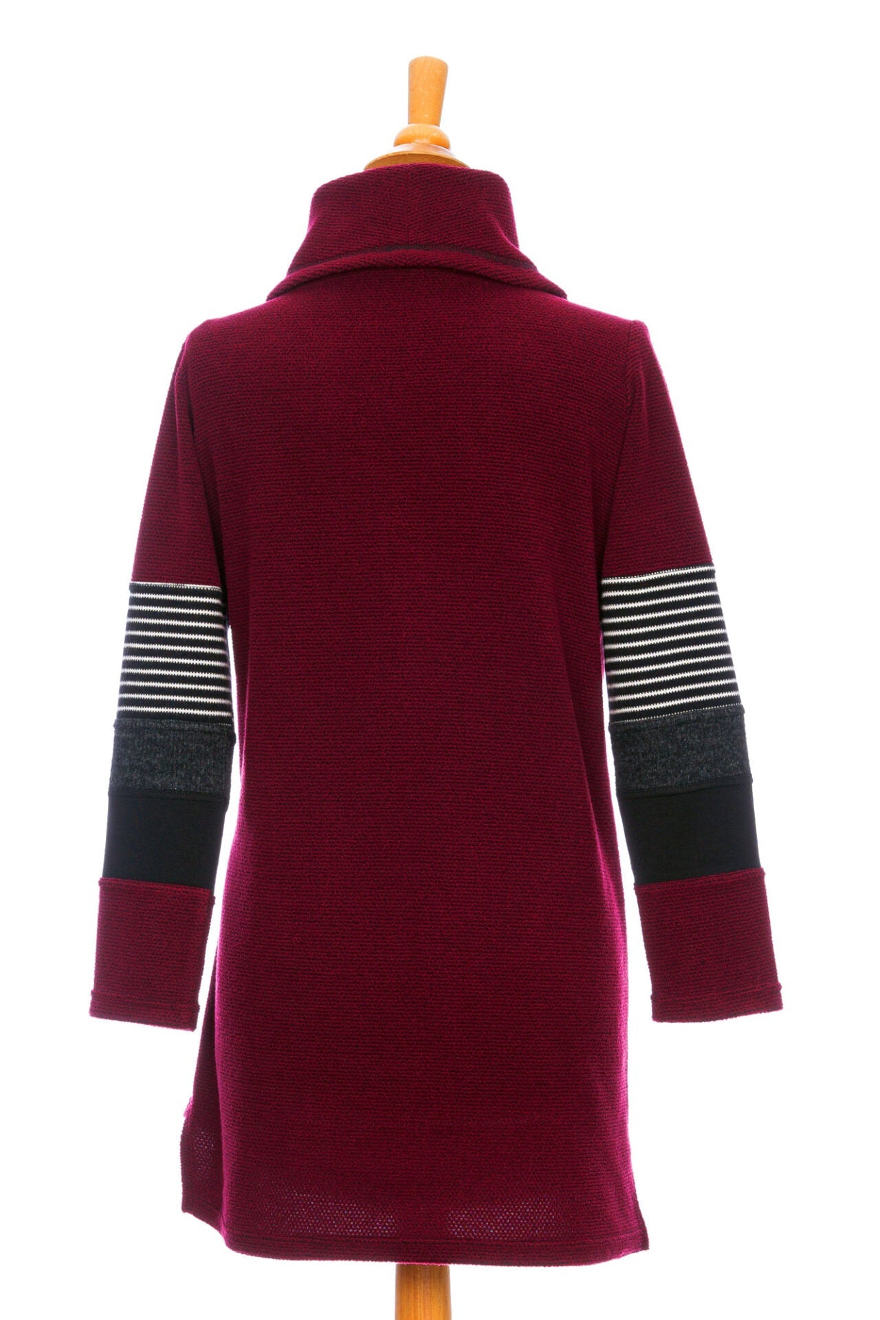Back view of the Églantier Tunic by Rien ne se Perd in textured Bordeaux fabric with striped and black colour-blocking on the long sleeves shown on a mannequin in front of a white background. It has a large turtleneck, a faux leather floral decal, and a side pocket. She is wearing it with black leggings and standing in a kitchen. 