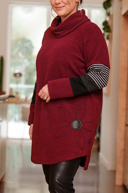 Side view of a woman wearing the Églantier Tunic by Rien ne se Perd in textured Bordeaux fabric with striped and black colour-blocking on the long sleeves. It has a large turtleneck, a faux leather floral decal, and a side pocket. She is wearing it with black leggings and standing in a kitchen. 