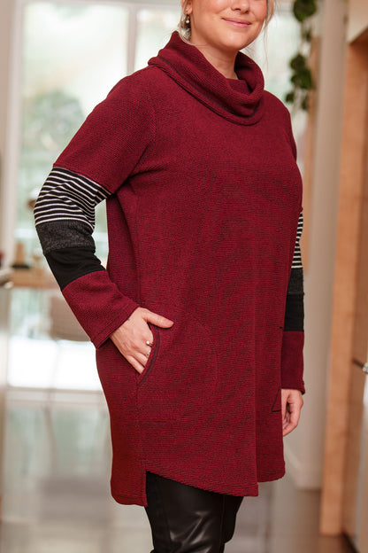 Side view of a woman wearing the Églantier Tunic by Rien ne se Perd in textured Bordeaux fabric with striped and black colour-blocking on the long sleeves. It has a large turtleneck, a faux leather floral decal, and a side pocket. She is wearing it with black leggings and standing in a kitchen. 