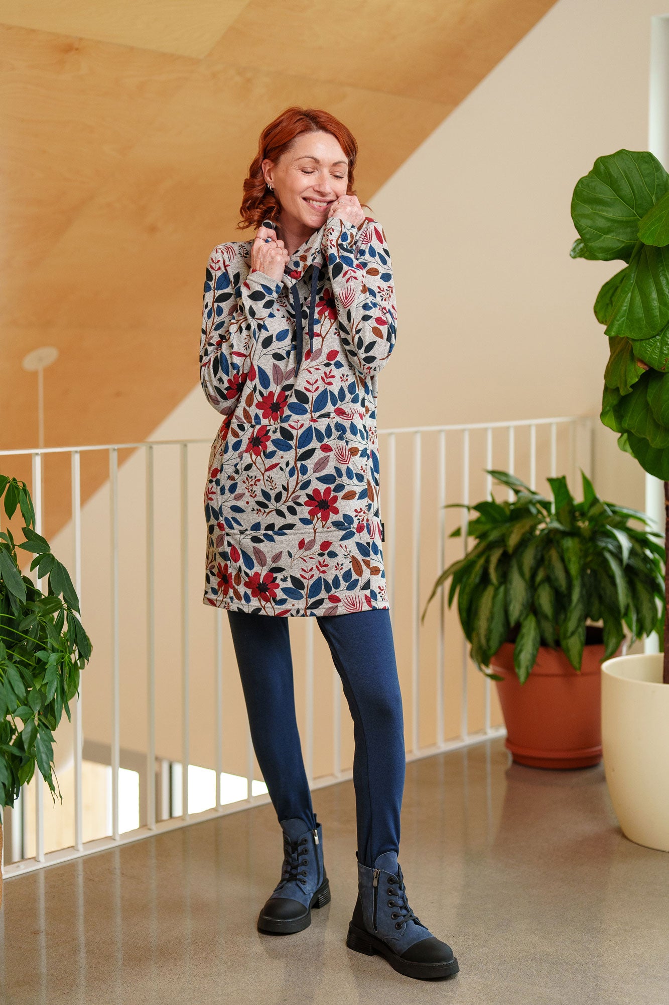 A woman wearing the Genevrier Tunic by Rien ne se Perd in Floral, featuring a turtleneck with a drawstring, a kangaroo pocket, long sleeves, and a straight cut. She is wearing it with navy leggings and standing indoors with a plant behind her. 