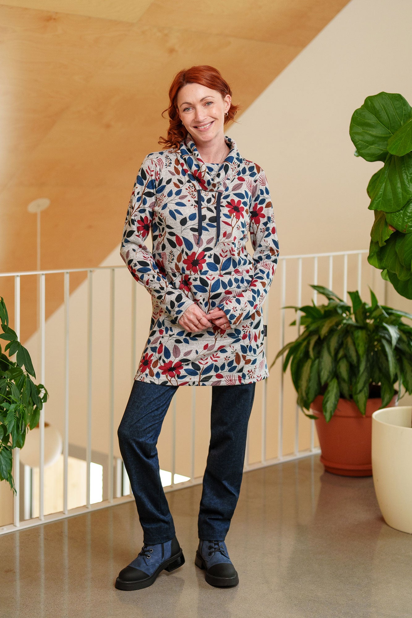 A woman wearing the Genevrier Tunic by Rien ne se Perd in Floral, featuring a turtleneck with a drawstring, a kangaroo pocket, long sleeves, and a straight cut. She is wearing it with navy leggings and standing indoors with  plants behind her. 