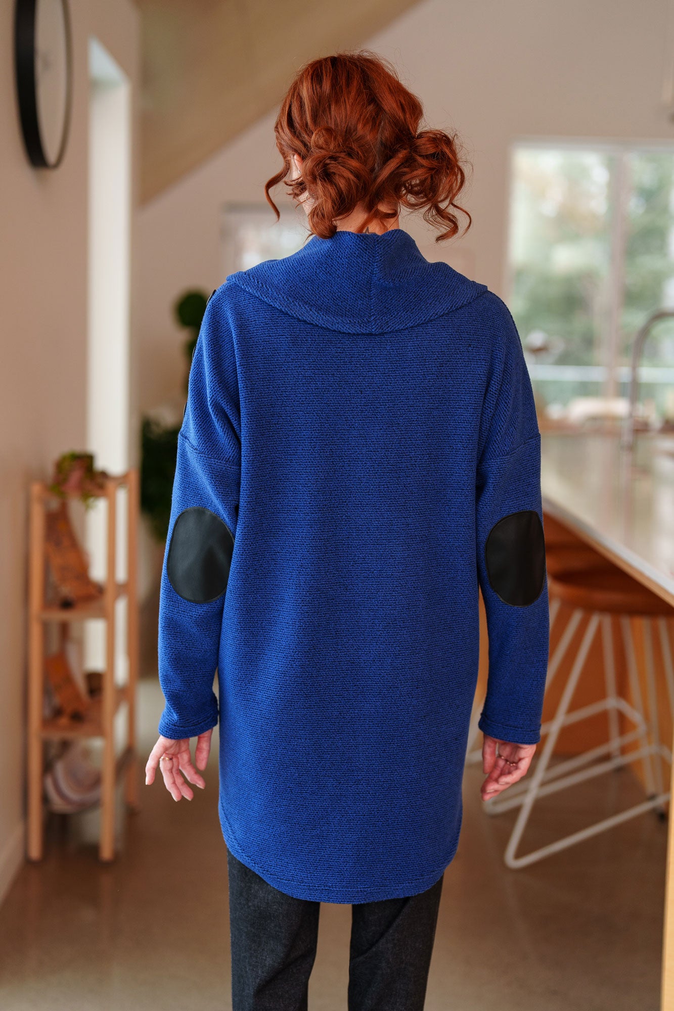 Back view of a woman wearing the Myrique Tunic by Rien ne se Perd in Blue, an oversized chenille tunic with a large cowl neck, rounded hem, faux leather elbow patches, button placket at the left shoulder, and patch pocket on the front. She is wearing it with the Resinosa Pants and standing in a kitchen. 