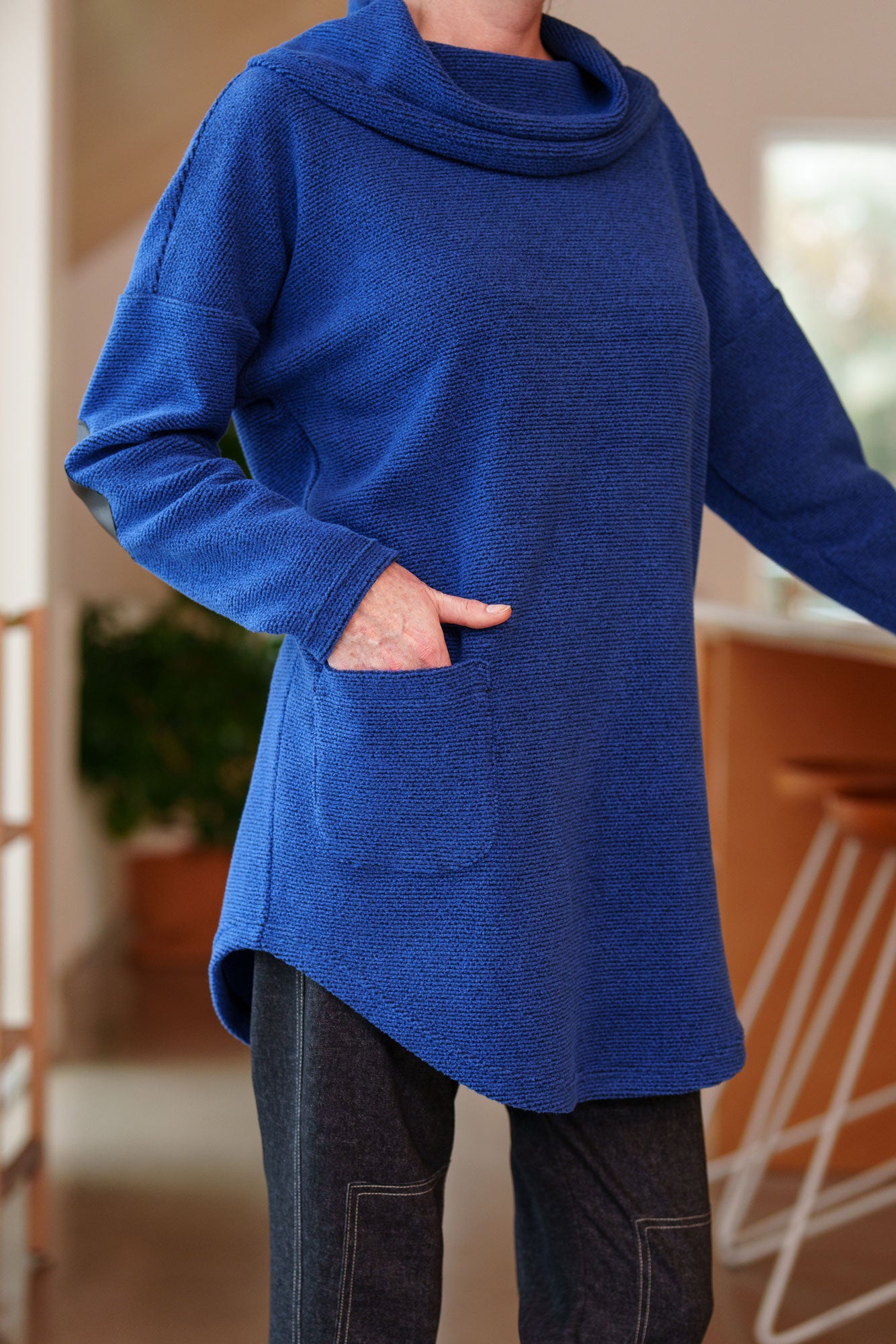 Close up view of a woman wearing the Myrique Tunic by Rien ne se Perd in Blue, an oversized chenille tunic with a large cowl neck, rounded hem, faux leather elbow patches, button placket at the left shoulder, and patch pocket on the front. She is wearing it with leggings and standing in a kitchen. 