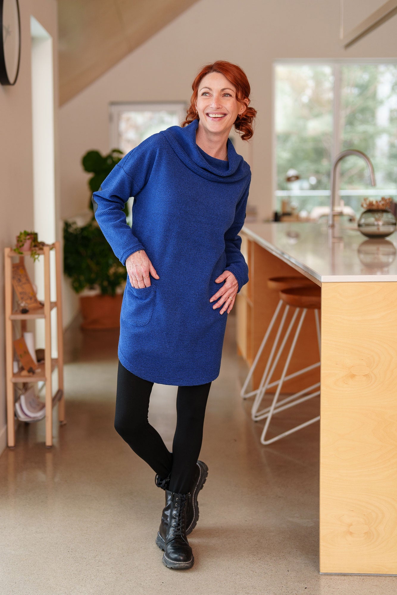 A woman wearing the Myrique Tunic by Rien ne se Perd in Blue, an oversized chenille tunic with a large cowl neck, rounded hem, faux leather elbow patches, button placket at the left shoulder, and patch pocket on the front. She is wearing it with leggings and standing in a kitchen. 