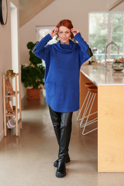 A woman wearing the Resinosa Pants by Rien ne se Perd in Black, slim fitting pants with faux leather at the front and plain fabric at the back, with a wide pull on waistband. She is wearing them with the Myrique Tunic in Blue and standing in a kitchen.
