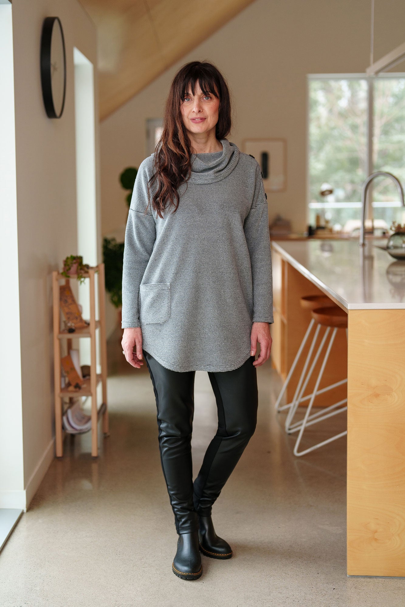 A woman wearing the Myrique Tunic by Rien ne se Perd in Grey, an oversized chenille tunic with a large cowl neck, rounded hem, faux leather elbow patches, button placket at the left shoulder, and patch pocket on the front. She is wearing it with the Resinosa Pants and standing in a kitchen. 