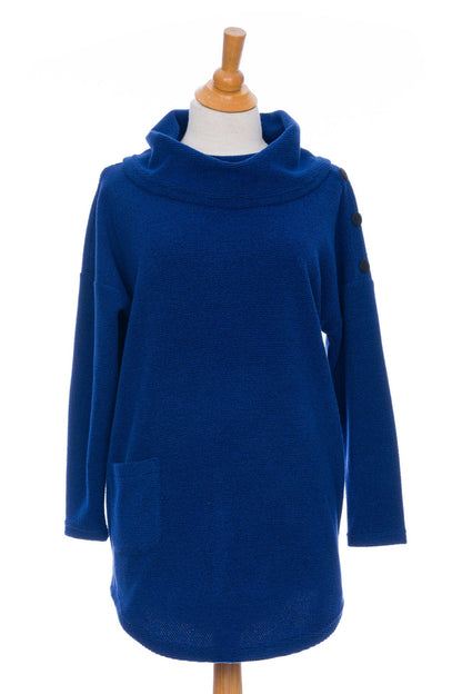 The Myrique Tunic by Rien ne se Perd in Blue, an oversized chenille tunic with a large cowl neck, rounded hem, faux leather elbow patches, button placket at the left shoulder, and patch pocket on the front, is shown on a mannequin in front of a white background. 