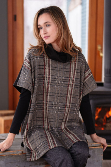 A woman wearing the Borduas Tunic-Poncho by Rien ne se Perd in a checkered print, sitting in front of a wood stove in a cabin.