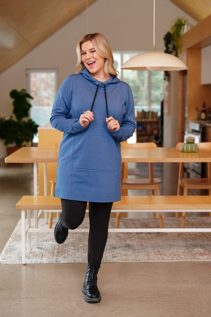 A woman wearing the Toundra Tunic by Rien ne se Perd in Blue, featuring a large hood with drawstrings, a kangaroo pocket, and a relaxed fit. She is wearing it with black leggings and is standing in a kitchen. 

