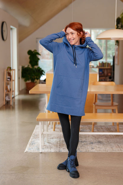 A woman wearing the Toundra Tunic by Rien ne se Perd in Blue, featuring a large hood with drawstrings, a kangaroo pocket, and a relaxed fit. She is wearing it with black leggings and is standing in a kitchen. 
