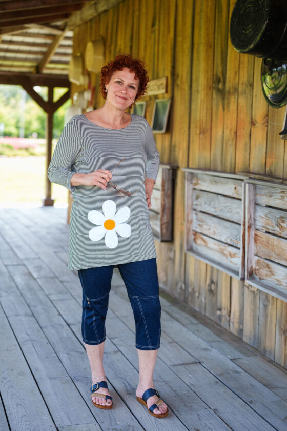 A woman wearing the LIndy Capri pants from Rien ne se Perd, faux denim stretch capris with a wide pull-on waist, contrast seams at the bottom, and decorative coconut buttons. She is wearing them with the Zumba Tunic and standing outside a wooden building. 