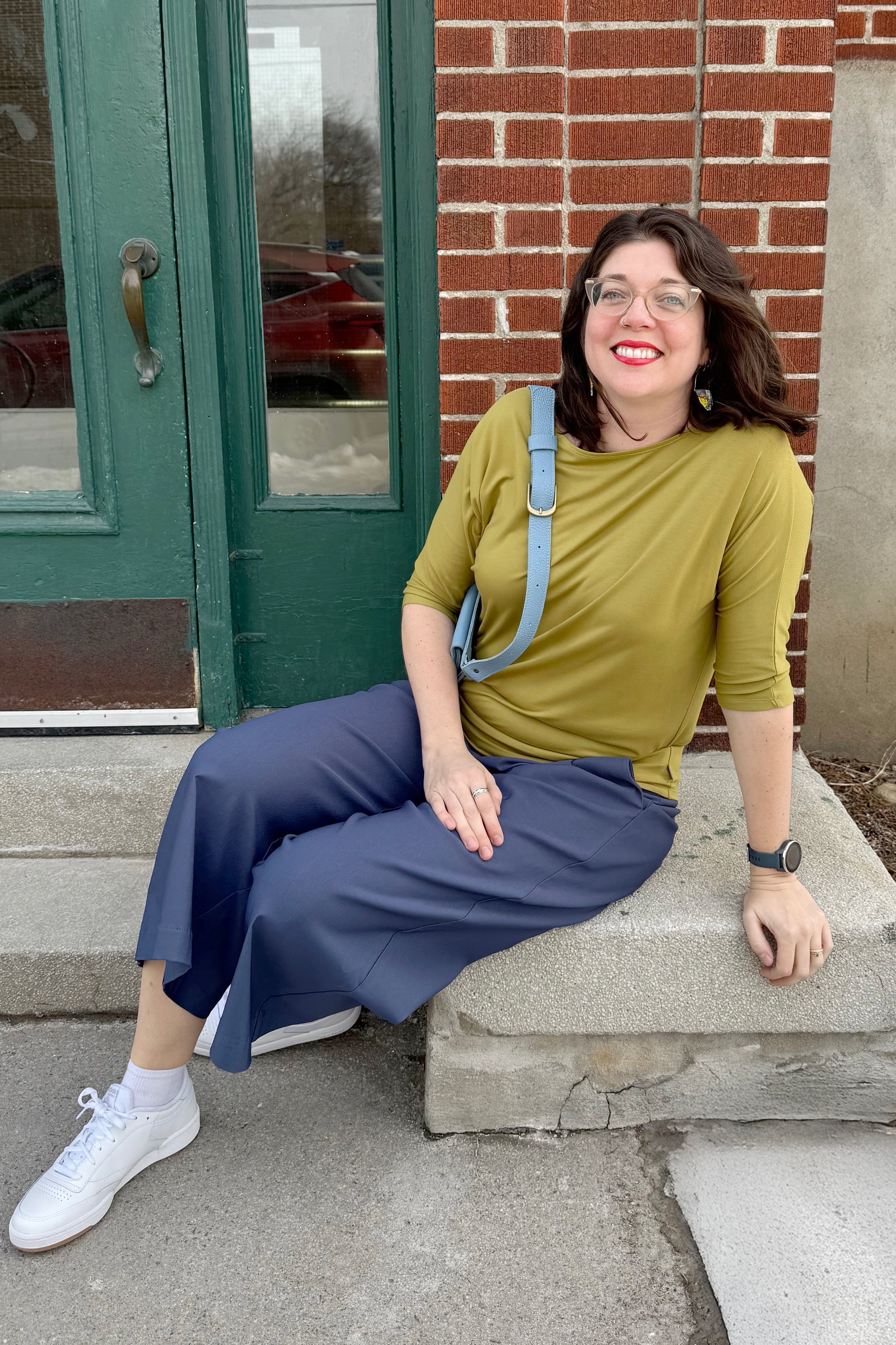 Woman sitting on steps wearing the Bettina Top by Moovment and blue pants, with a brick building in the background.