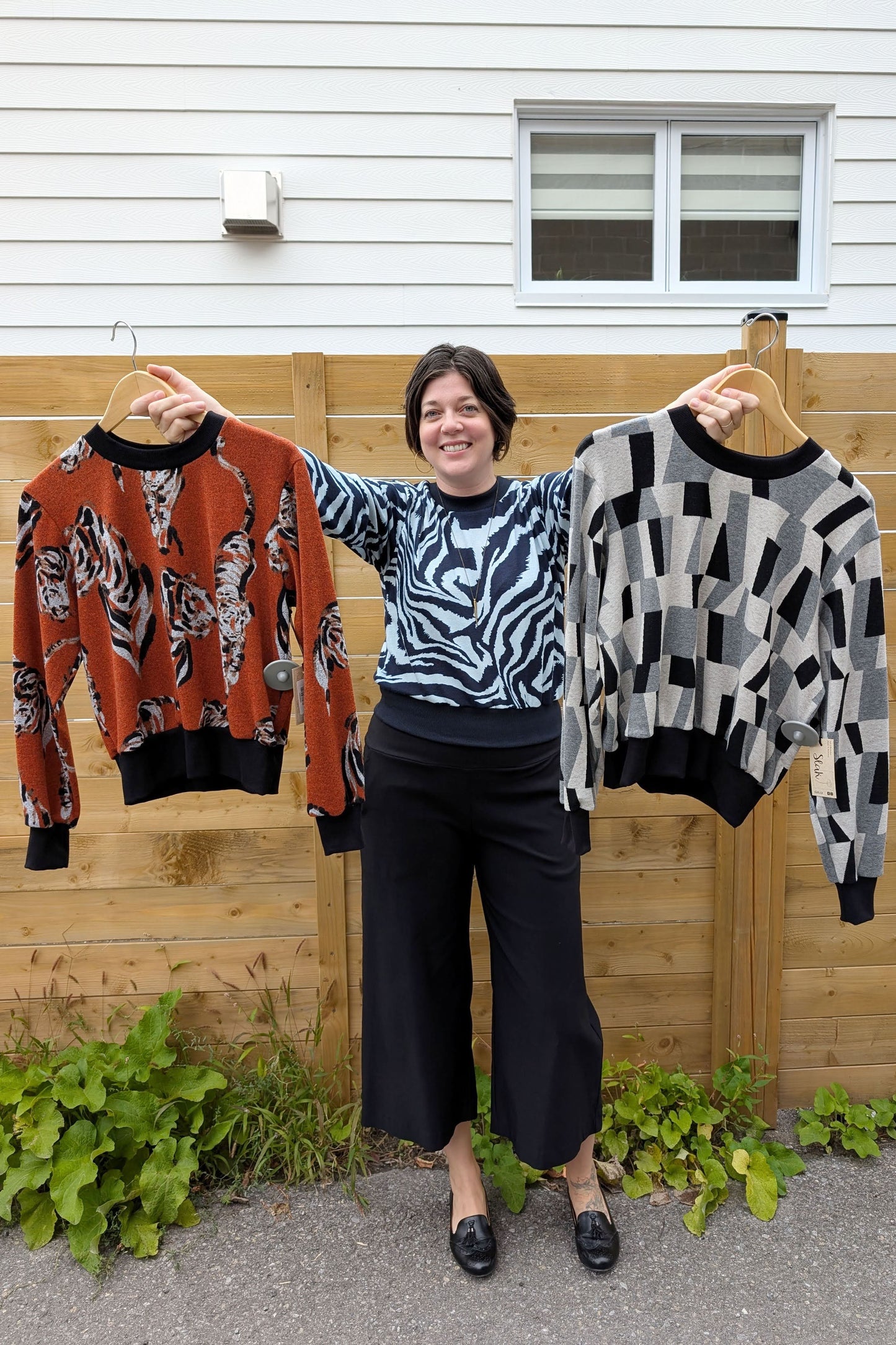 A woman wearing the Wanda Sweater in Blue Zebra print by Slak with the Mabel Pants in Black by Studio D, holding up the Wanda Sweater in Tiger and Black with Grey Prints, standing outside in front of a wooden fence