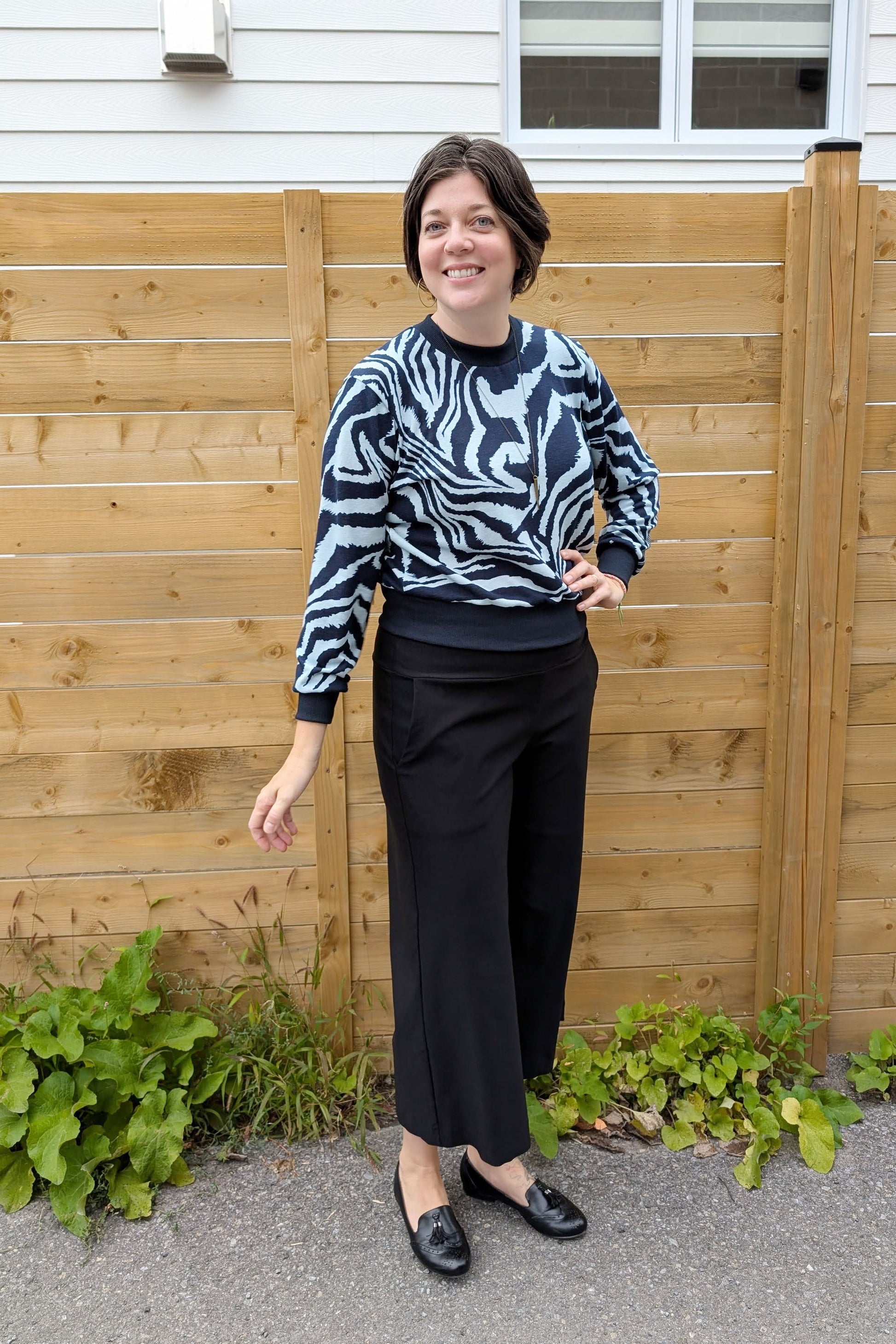 A woman wearing the Wanda Sweater in Blue Zebra print by Slak with the Mabel Pants in Black by Studio D, standing outside in front of a wooden fence