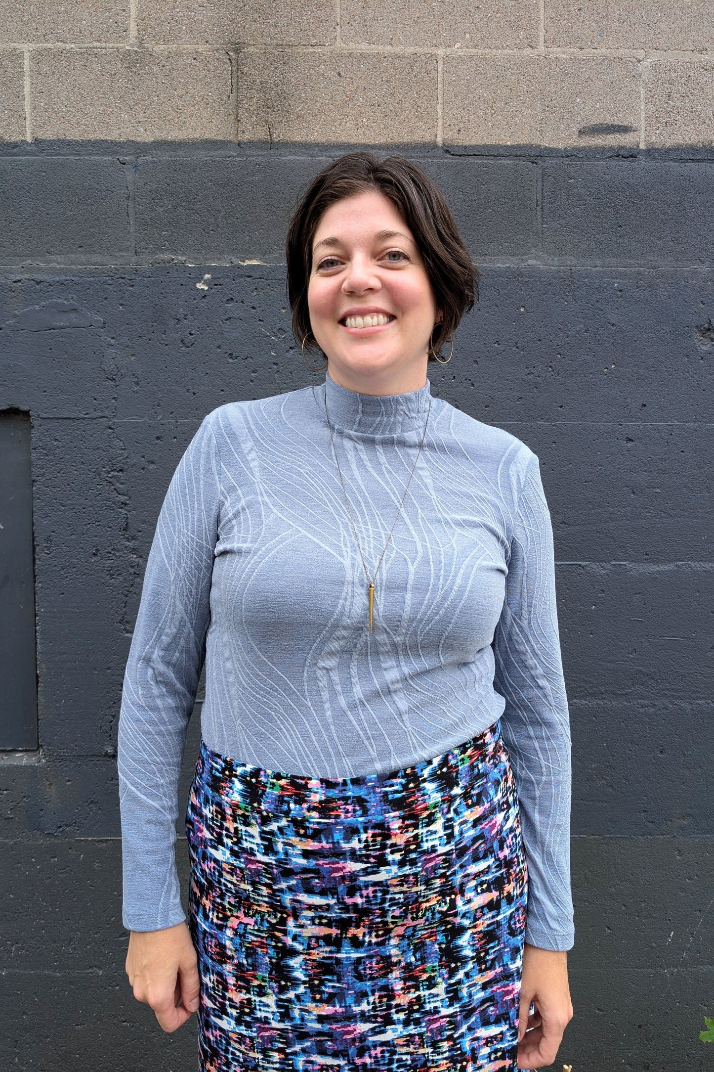 A woman wearing the Franco Mock Top by Luc Fontaine in blue, featuring a textured fabric, mock turtleneck and long sleeves, with the Eve Skirt by Slak, standing outside in front of a brick wall. 