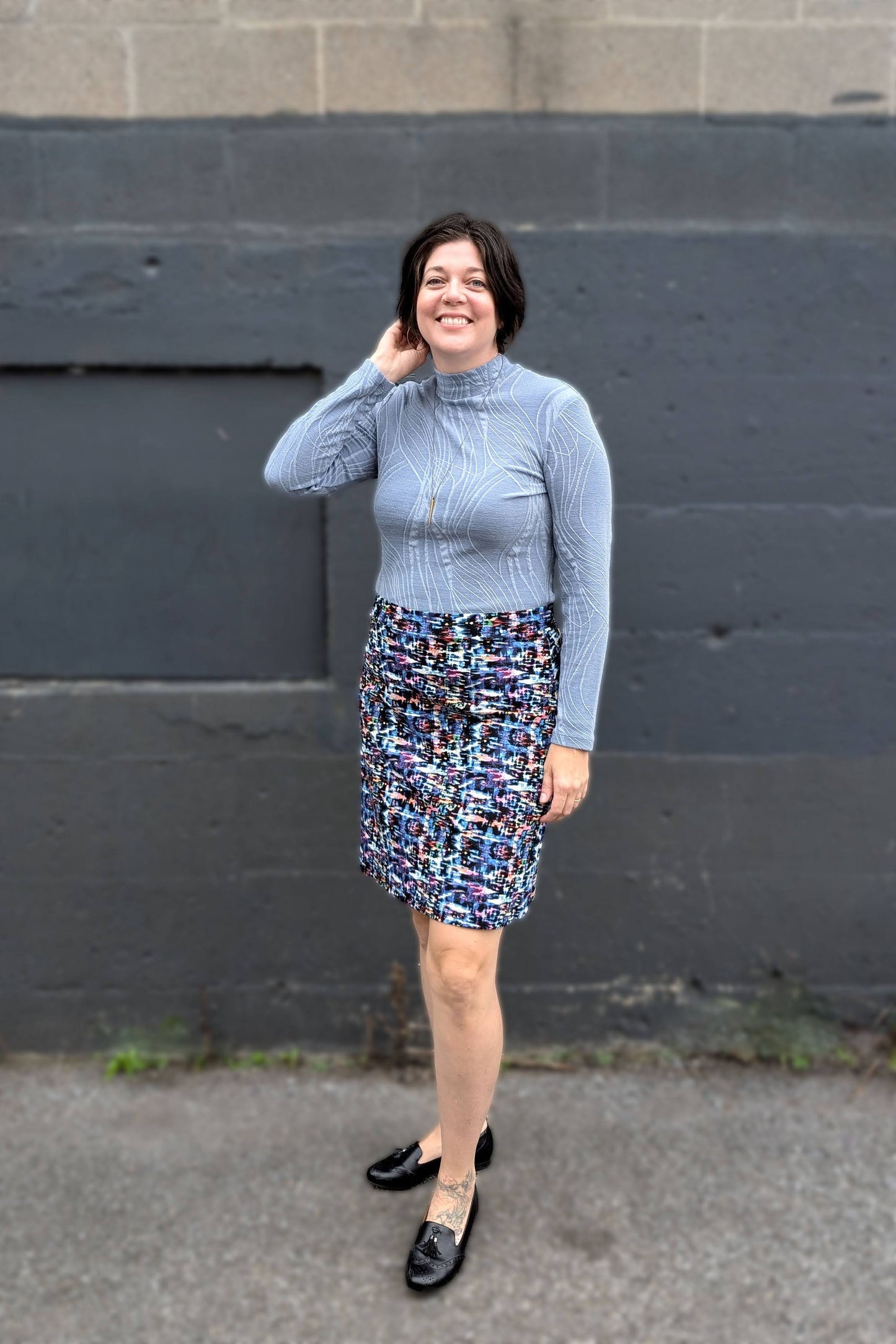 A woman wearing the Franco Mock Top by Luc Fontaine in blue, featuring a textured fabric, mock turtleneck and long sleeves, with the Eve Skirt by Slak, standing outside in front of a brick wall. 