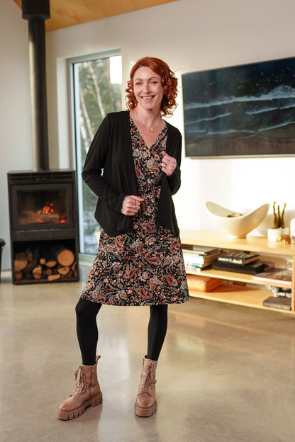 A woman wearing the Epilobe Dress by Rien ne se Perd in Fall Paisley, a classic wrap dress with a tie at the waist, delicate pleats at the shoulders, and a hem that hits above the knee. She is wearing a black cardigan over it and is standing in a living room with a fireplace and TV.