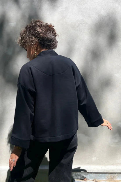 Back view of a woman wearing the Thuya Jacket by Kazak in Black, standing in front of a grey wall 