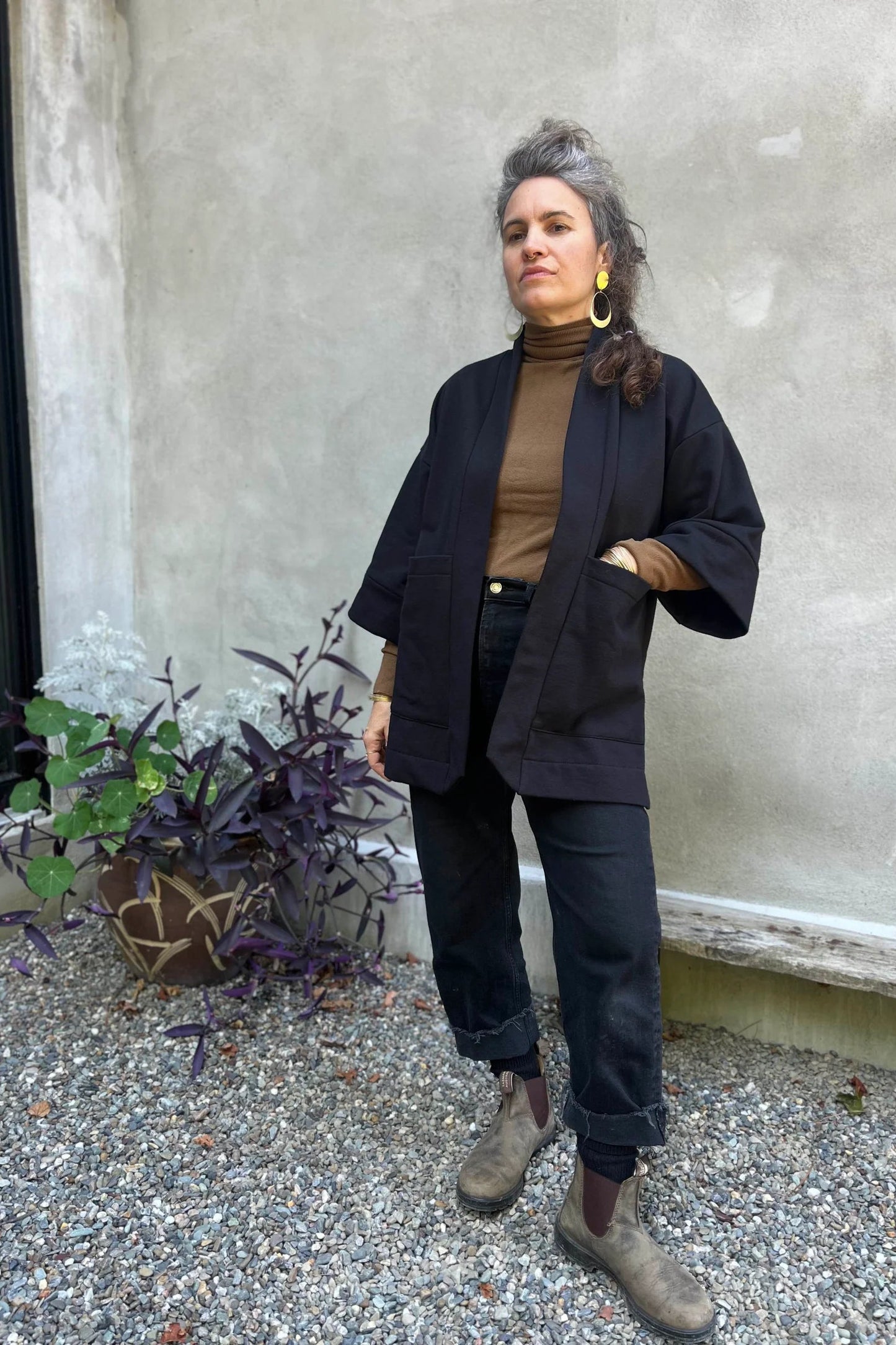 A woman wearing the Cydonia Cardigan by Kazak in Black, a hip-length open cardigan with 3/4 kimono sleeves and large patch pockets. She is wearing it with black pants and a black top and standing outside.