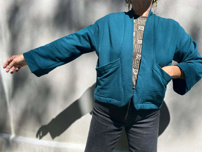 A woman wearing the Thuya Jacket by Kazak in Teal, standing in front of a grey wall 