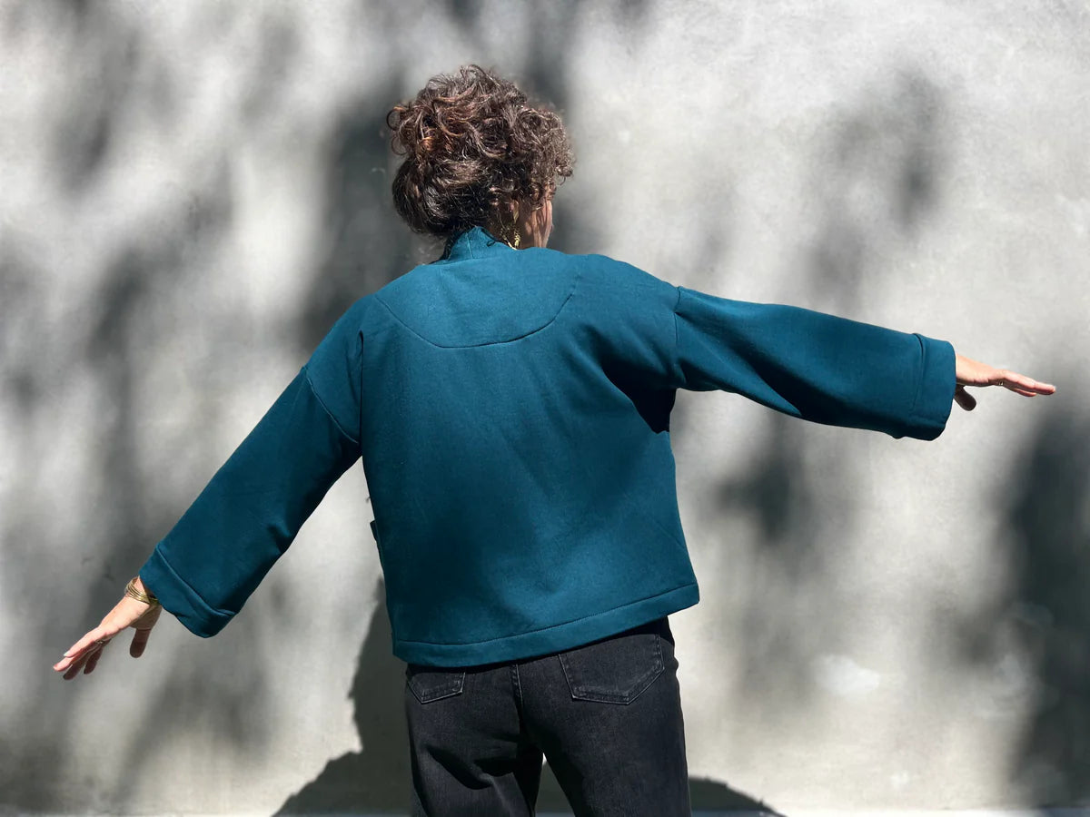 Back view of a woman wearing the Thuya Jacket by Kazak in Tea;, standing in front of a grey wall 