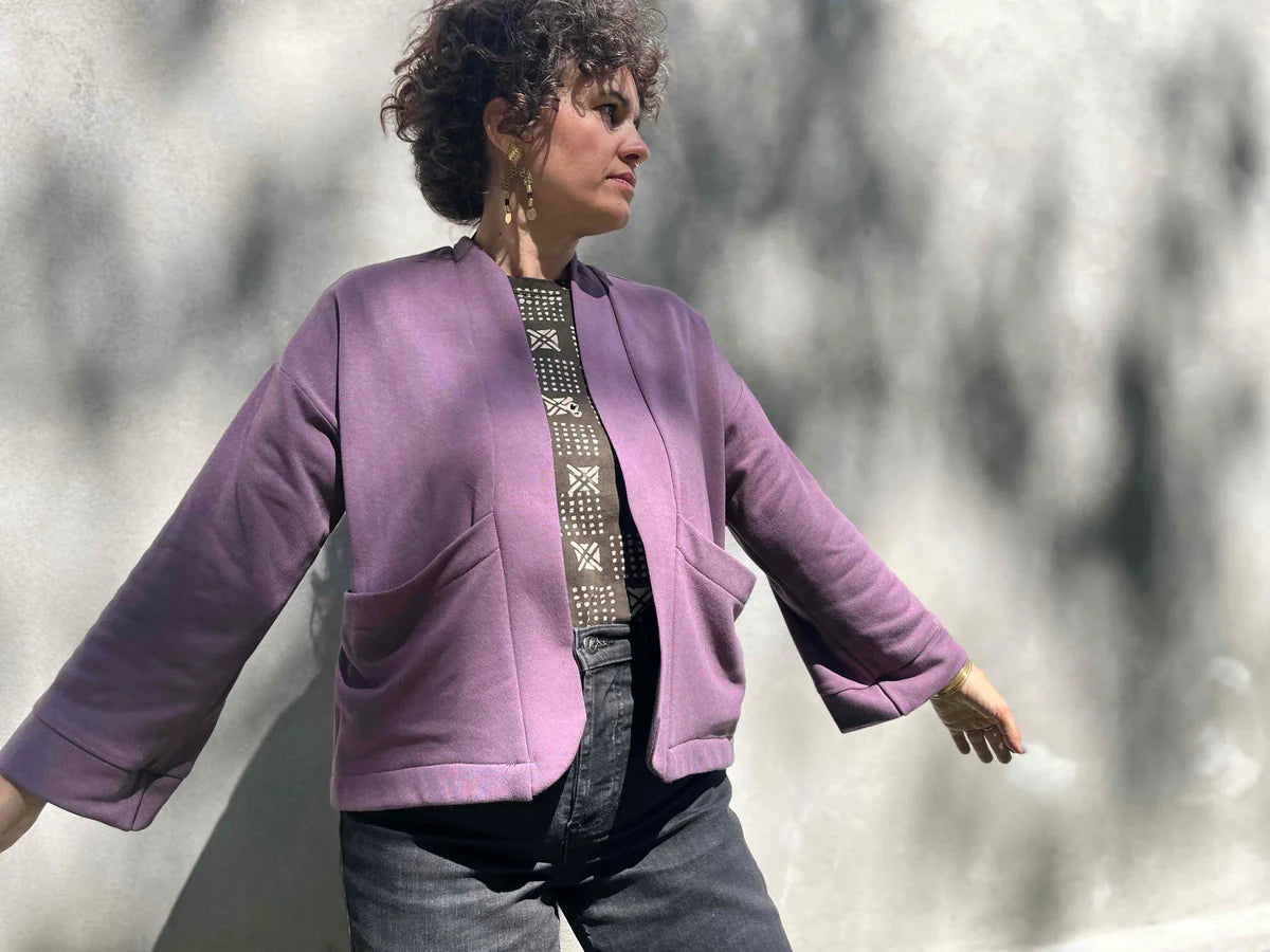 A woman wearing the Thuya Jacket by Kazak in Purple Haze, standing in front of a grey wall 