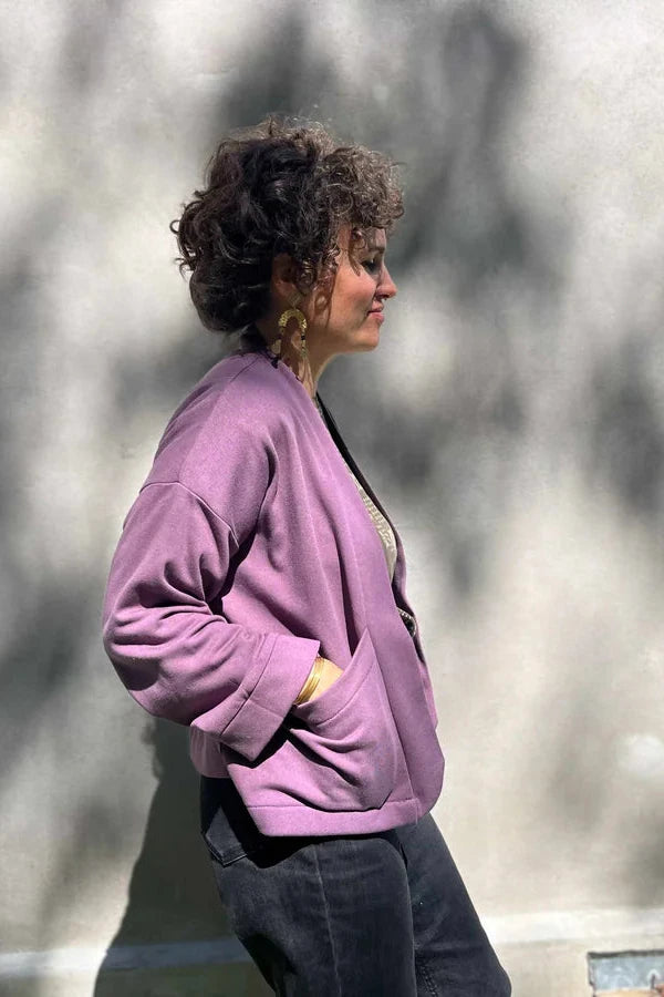 Side view of a woman wearing the Thuya Jacket by Kazak in Purple Haze, standing in front of a grey wall 