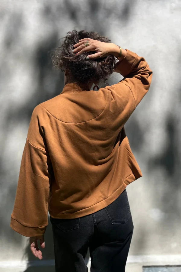 Back view of a woman wearing the Thuya Jacket by Kazak in Teak, standing in front of a grey wall 