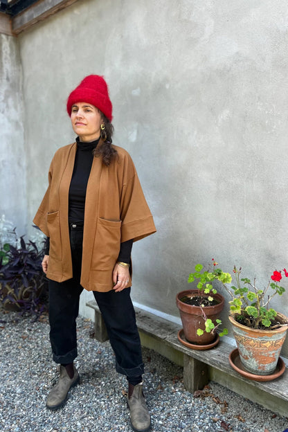 A woman wearing the Cydonia Cardigan by Kazak in Teak, a hip-length open cardigan with 3/4 kimono sleeves and large patch pockets. She is wearing it with black pants and a black top and standing outside.