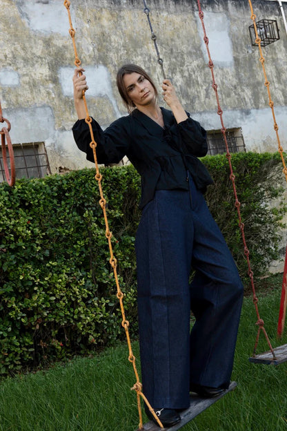 A woman wearing the Vienna Pants in Jeans by MAS, high waisted pants with wide legs and a zipper and tab closure. She is wearing them with a black top and standing on a swing.