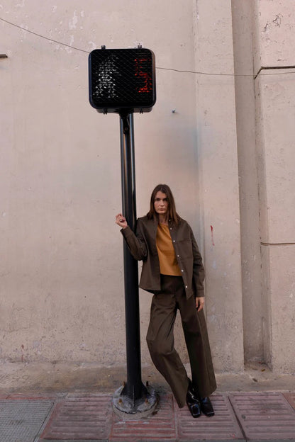 A woman wearing the Vienna Pants in Capers by MAS, high waisted pants with wide legs and a zipper and tab closure. She is wearing them with yellow top and matching jacket and standing on the street. 