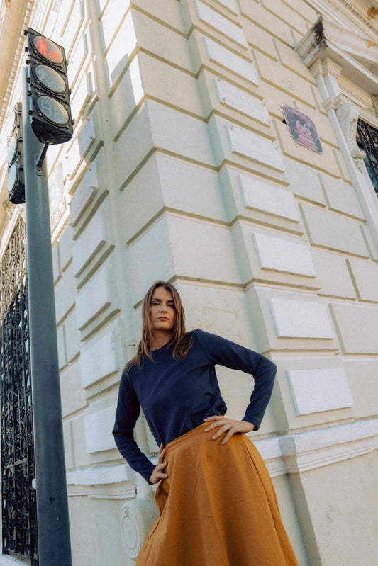 A woman wearing the Whistler Long Sleeve Top in Blueberry, a ribbed cotton knit with a round neck and straight cut. She is wearing it with a  brown skirt and standing against a light-colored building.