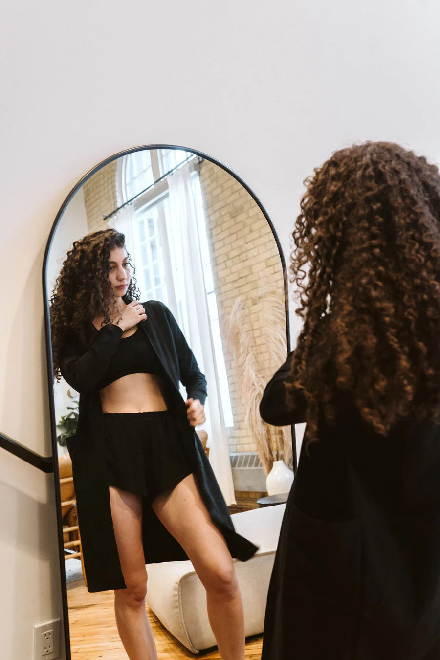 A woman wearing the Courtney Cardigan by Advika in Black, a long pen cardigan with patch pockets, made from tencel/organic eco-fabric, over shorts and a bra. She is looking at her reflection in a mirror. 