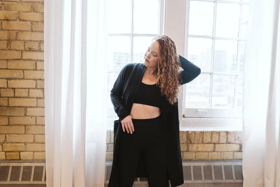 A woman wearing the Courtney Cardigan by Advika in Black, a long pen cardigan with patch pockets, made from tencel/organic eco-fabric, over legggings and a bra. She is standing in front of a window in a brick wall. 