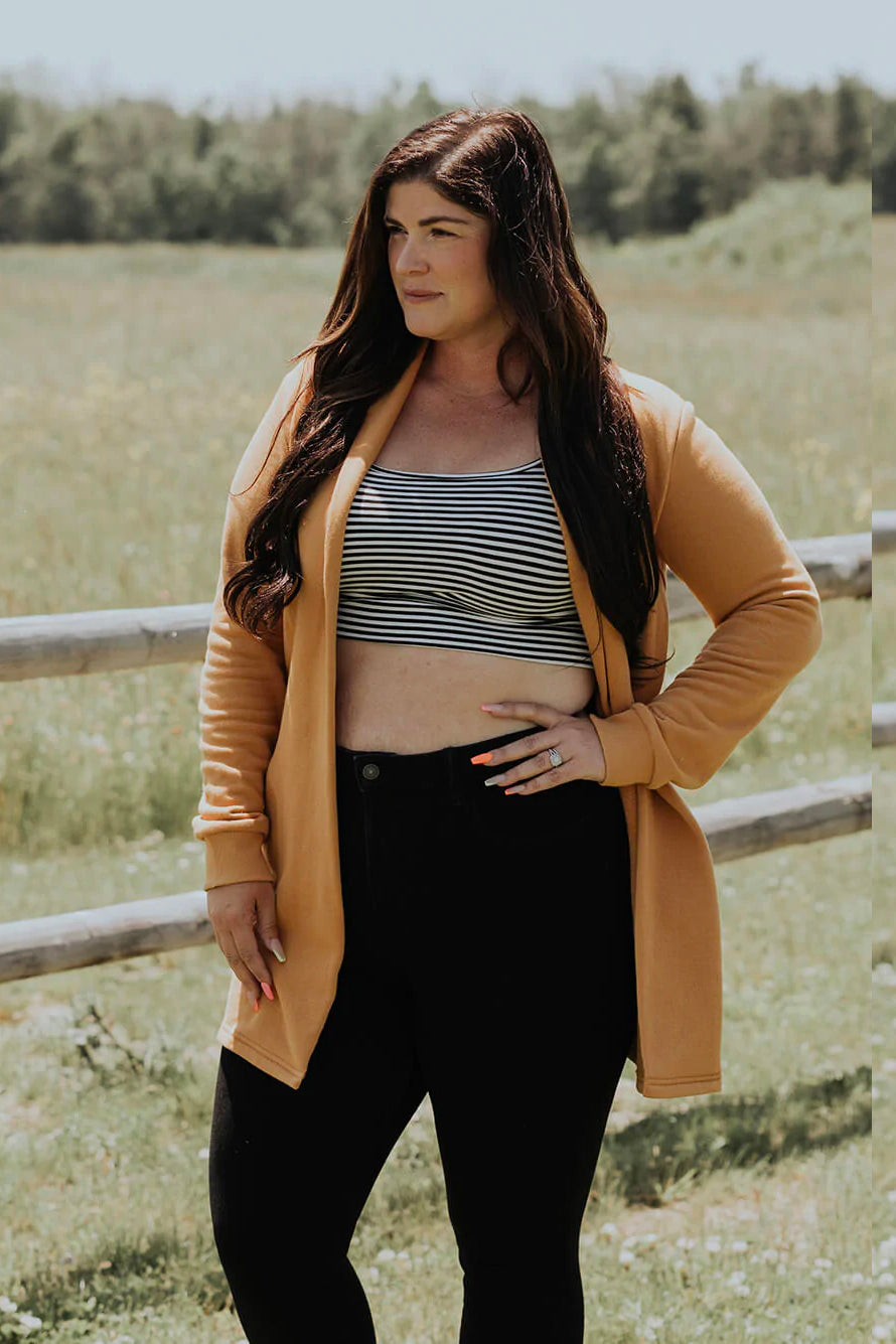 A woman wearing the Classic Briton Cardigan by Blondie Apparel in Gold with black jeans and a striped crop top, standing in front of a fence and a field