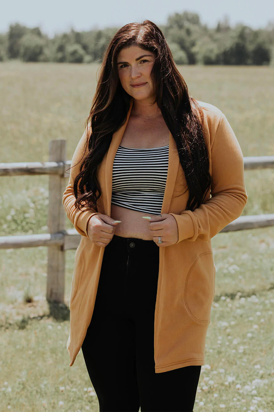 A woman wearing the Classic Briton Cardigan by Blondie Apparel in Gold with black jeans and a striped crop top, standing in front of a fence and a field